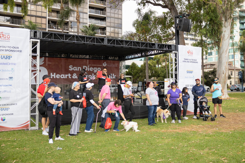 A group of people with dogs and a baby gather near a stage at the "San Diego Heart & Stroke Walk" event. Some are holding leashes, and others stand nearby, enjoying the outdoor gathering with trees and buildings in the background.