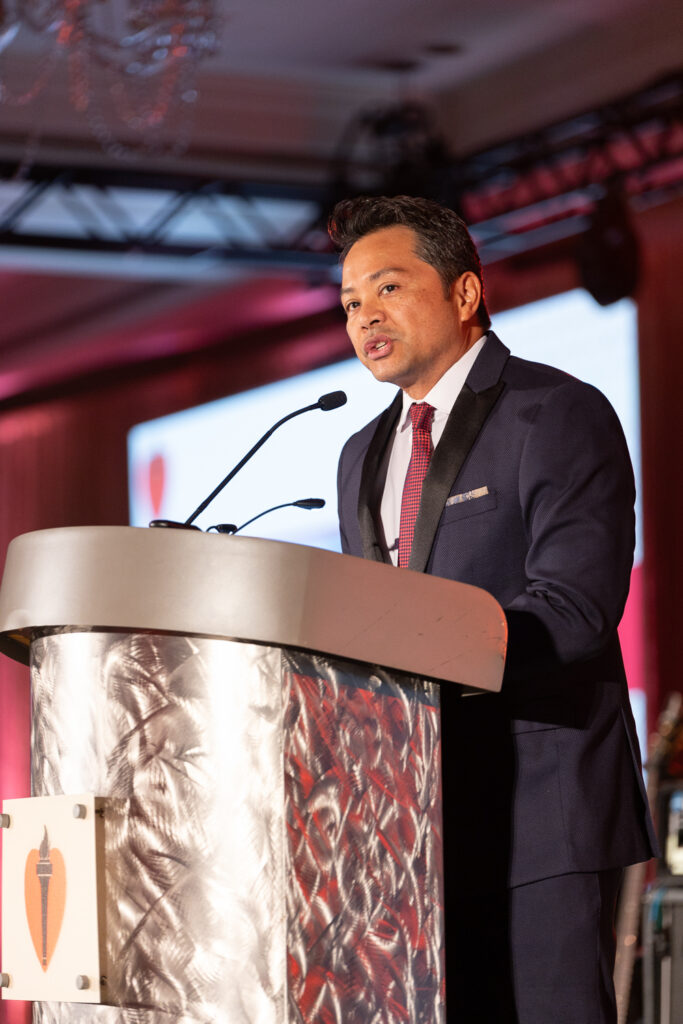 A man in a suit and tie speaks at a podium with microphones. The podium displays a heart emblem. The background features a large screen and is lit with red lighting.