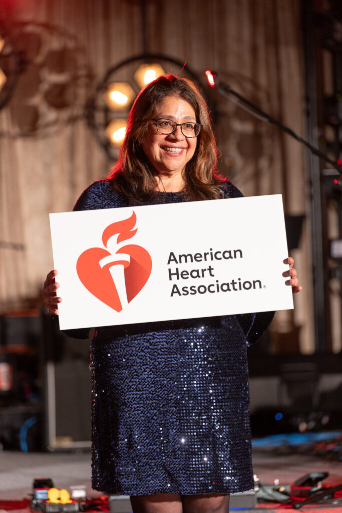 A woman in a sequined dress holds a sign with the American Heart Association logo. She stands on a stage with warm lighting in the background.