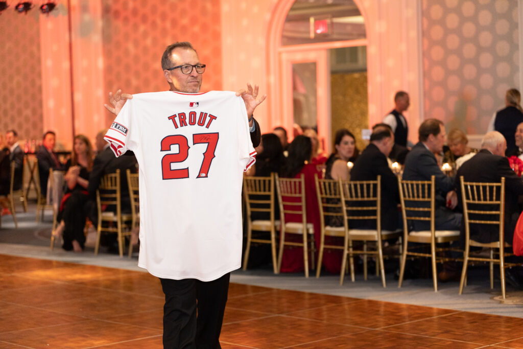 A man in glasses holds up a white sports jersey with "TROUT" and "27" printed on it. The event is indoors with seated guests at round tables in the background. The atmosphere seems formal with dim lighting.