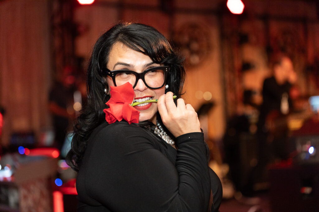 A person with long dark hair and glasses holds a red rose in their mouth, wearing a black top and smiling playfully. The background is softly lit with warm lights and out-of-focus silhouettes of people and musical equipment.