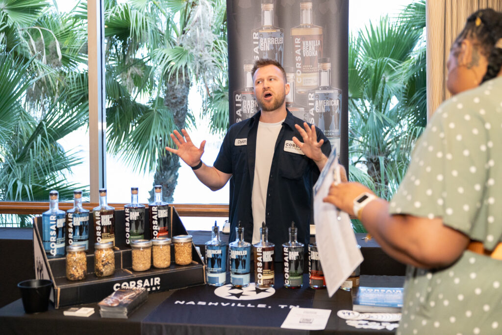 A man stands behind a display table with Corsair-branded bottles and promotional materials. He is gesturing with his hands while speaking to a woman holding a pamphlet. In the background are tall windows and lush greenery.