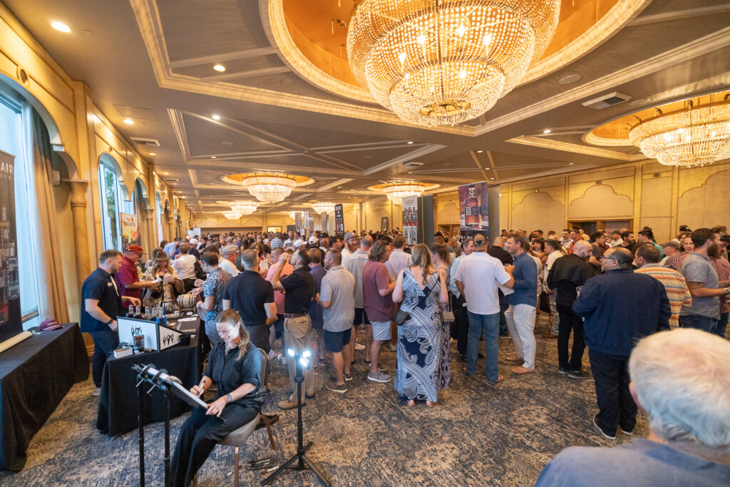 A large crowd of people mingling in an elegant ballroom with ornate chandeliers and high ceilings. A musician is playing a keyboard on the left, while attendees gather around various displays and exhibits.