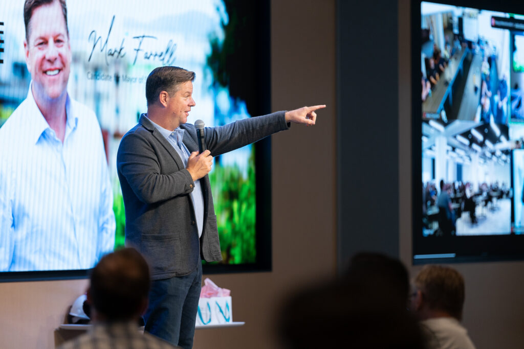 A man in a blue suit holds a microphone and points while speaking on stage. Behind him is a large screen displaying his image and text. Another screen shows an audience seated in a conference room.