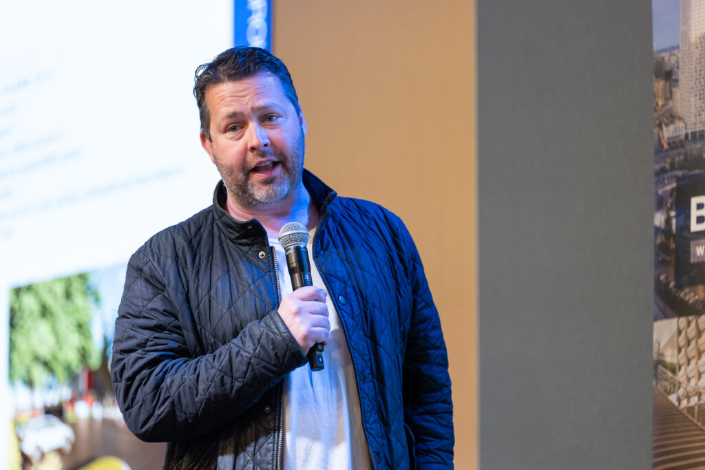 A man wearing a dark quilted jacket speaks into a microphone. He stands in front of a screen displaying a presentation slide, with city scenery partially visible on the screen.