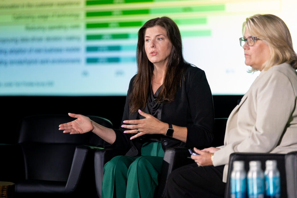 Two women are seated in armchairs on a stage, engaged in a discussion. One is speaking while gesturing with her hands, and the other listens attentively. A colorful bar graph is visible on the screen behind them. Bottled water is on a table nearby.