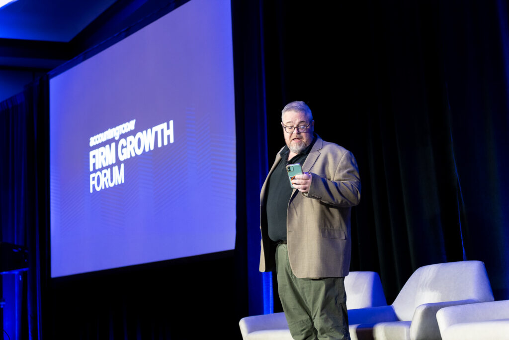 A person in a beige blazer stands on stage holding a phone, with "FIRM GROWTH FORUM" displayed on a large screen behind them. Two white chairs are on the stage.