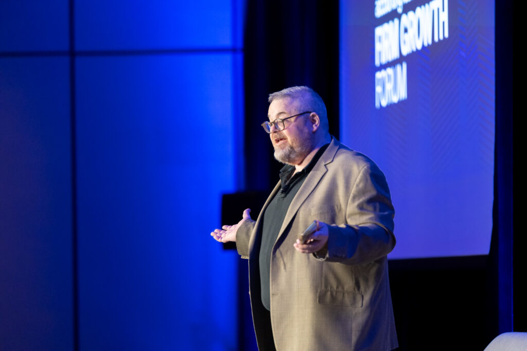 A man in a beige blazer and black shirt is speaking on stage at an event. He gestures with his hands while standing in front of a large screen displaying text. The background is illuminated with blue lighting.