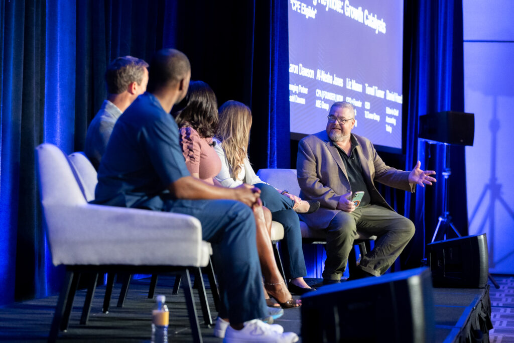 A panel of five people sits on stage, engaged in a discussion at a conference. A large screen in the background displays text. One panelist gestures while speaking. The audience is not visible.