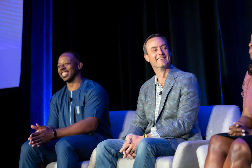 Two men are sitting on a stage panel, facing the audience. The man on the left wears a blue shirt, and the man on the right wears a plaid jacket. They both appear engaged and smiling. The background is dark with blue lighting.