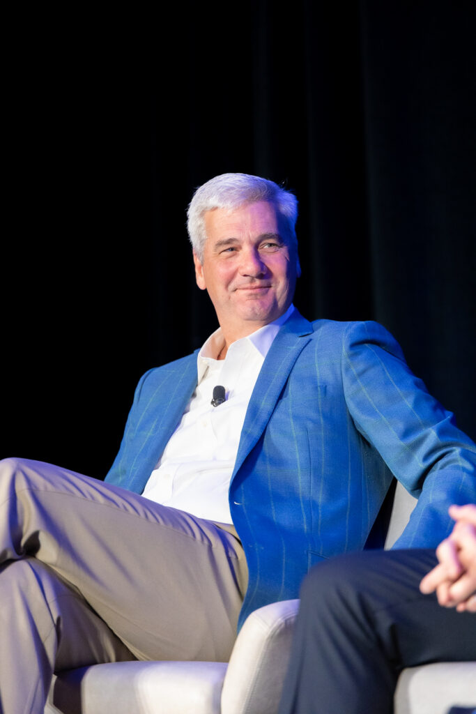 A person with gray hair wears a blue blazer and sits on a stage in a white chair. They are smiling and looking slightly to the right. A microphone is clipped to their shirt.