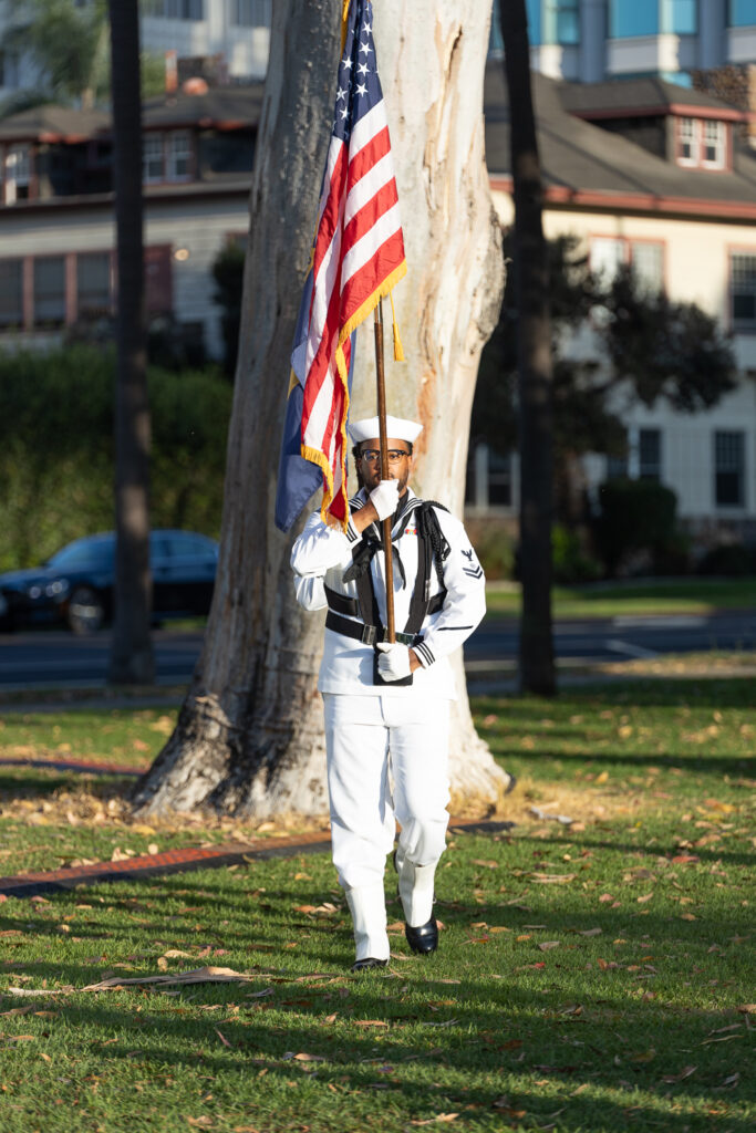 A person in a white military uniform carries the American flag with other flags behind them. They are walking on grass next to a large tree with buildings in the background.