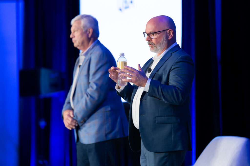 Two men in suits stand on a stage. The man on the right holds a bottle while speaking. The stage is lit with blue lights, and a screen is visible in the background.