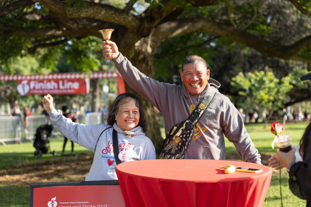 Two smiling people celebrate at a park event. The man holds a small bell. They stand by a red table with an American Heart Association logo. A "Finish Line" banner is visible in the background, along with trees and fencing.