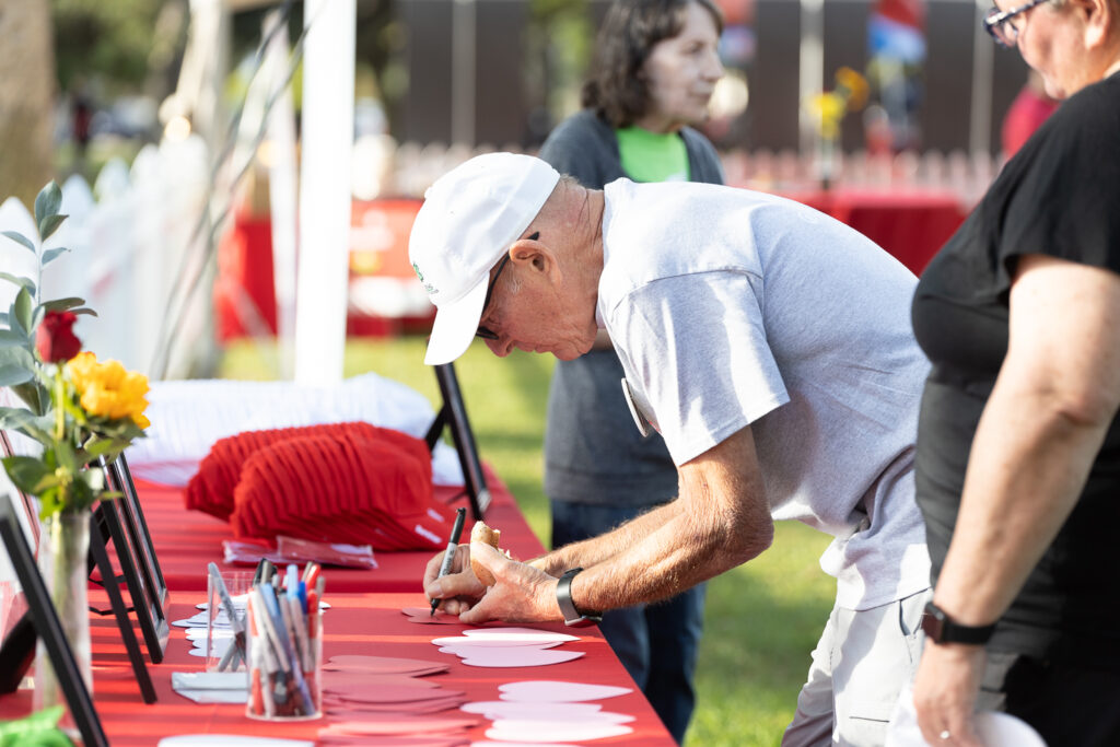 An elderly man wearing a white cap writes on a red heart-shaped card at an outdoor table. The table is covered with art supplies and framed photos. Other people are gathered around, and there are red tablecloths and flowers nearby.