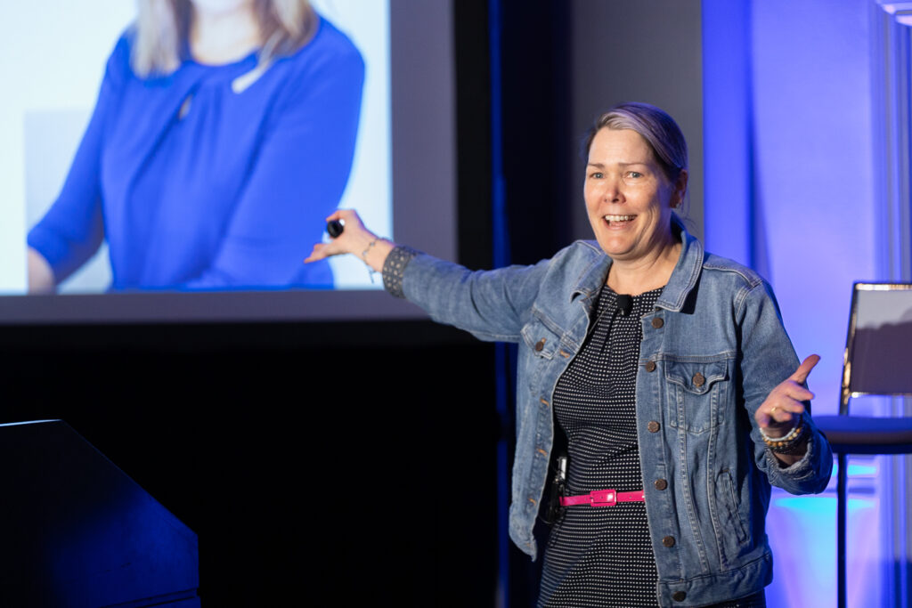 A woman wearing a denim jacket and a black dress is speaking energetically at a presentation. She is gesturing with one hand toward a large screen displaying an image of a woman in a blue top.