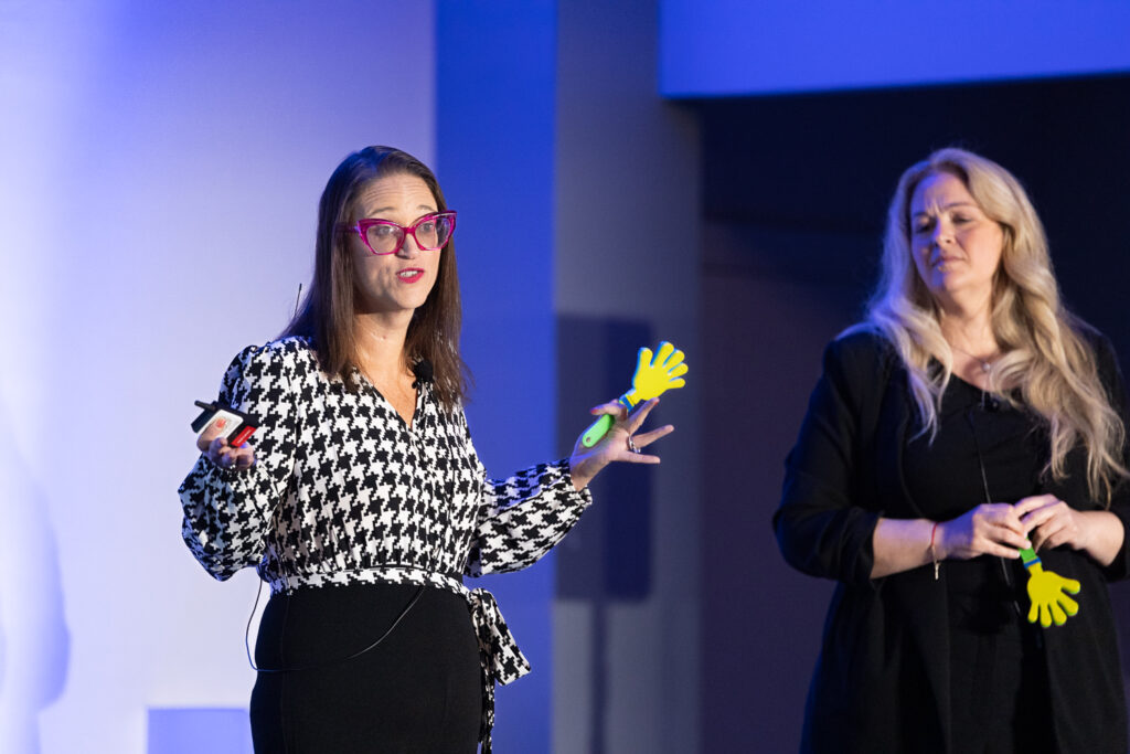 Two women are on stage speaking. The woman on the left is gesturing with colorful hand-shaped props, wearing a black and white houndstooth outfit and pink glasses. The woman on the right has long blonde hair and is wearing a black outfit.