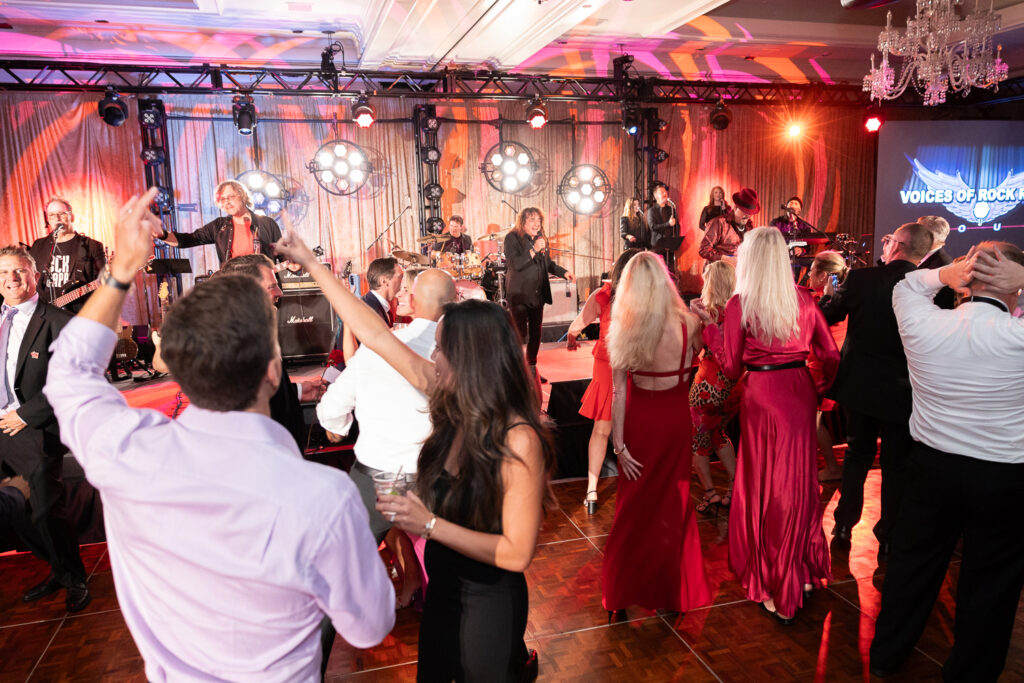 A lively crowd dances in front of a stage where a band is performing under bright lights. The event features a mix of formally dressed men and women, some with drinks in hand, while a colorful backdrop reads "Voices of Rock.