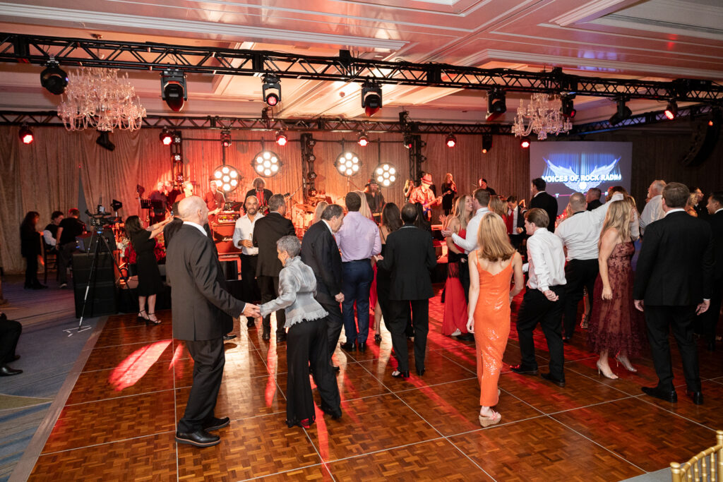 A lively dance floor scene at an indoor event. People of various ages are dancing and socializing under bright, colorful stage lights. A live band performs in the background, and a screen displays "Voices of Rock Radio.