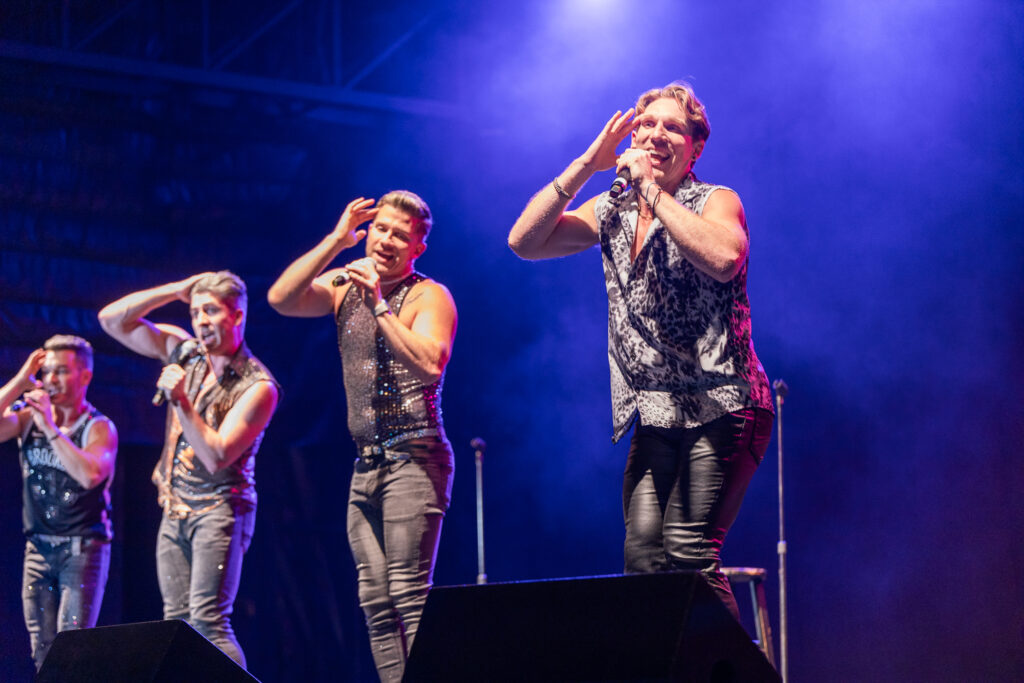 Five performers in shimmering outfits sing energetically on a dimly lit stage, each holding a microphone. They are animated, with vibrant expressions and dynamic poses, against a backdrop of deep blue lighting.