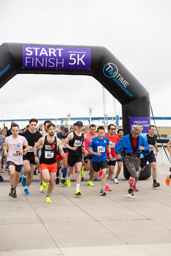 A group of runners starting a 5K race, passing under a black and purple inflatable arch that reads "Start Finish 5K." They are on a paved outdoor area, with a cloudy sky in the background.