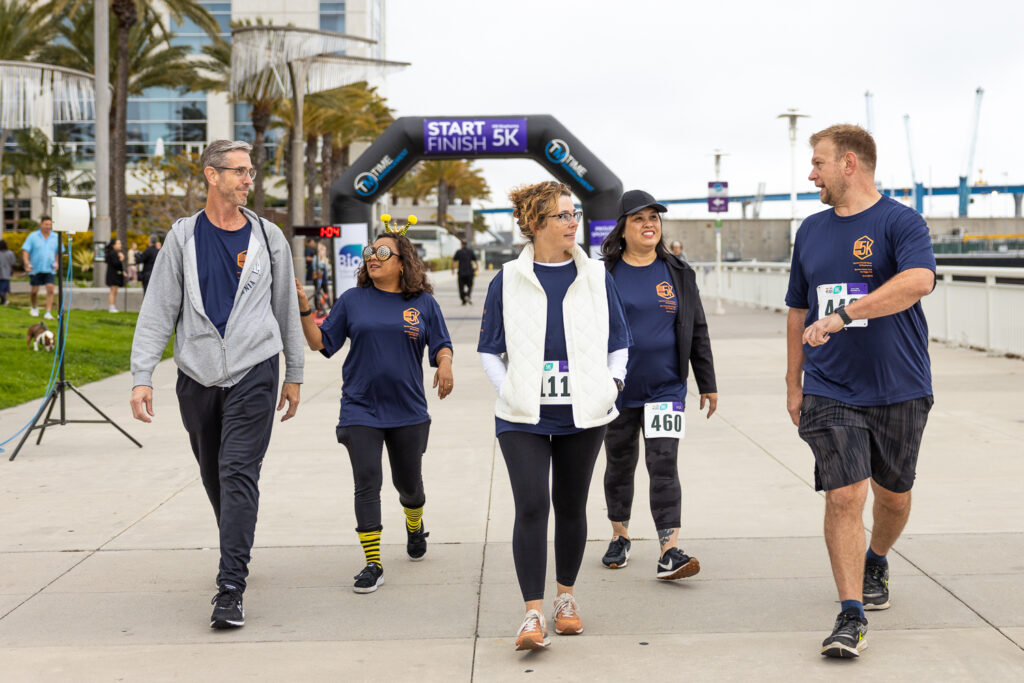 A group of five people in casual athletic wear walk together toward a 5K race finish line. They appear cheerful and are wearing numbered bibs. Palm trees and a waterfront are visible in the background.