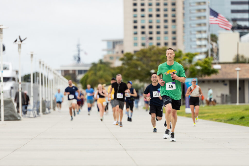 A group of people of various ages participating in a running event on a paved path. The front runner wears a green shirt with number 45. Buildings and an American flag are visible in the background.