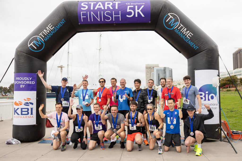 A group of runners poses under a "Start Finish 5K" arch. They display medals, wearing athletic gear, and smiling at the camera. Buildings and a cloudy sky are visible in the background. Signs for "KBI Biopharma" and another sponsor are on the arch.