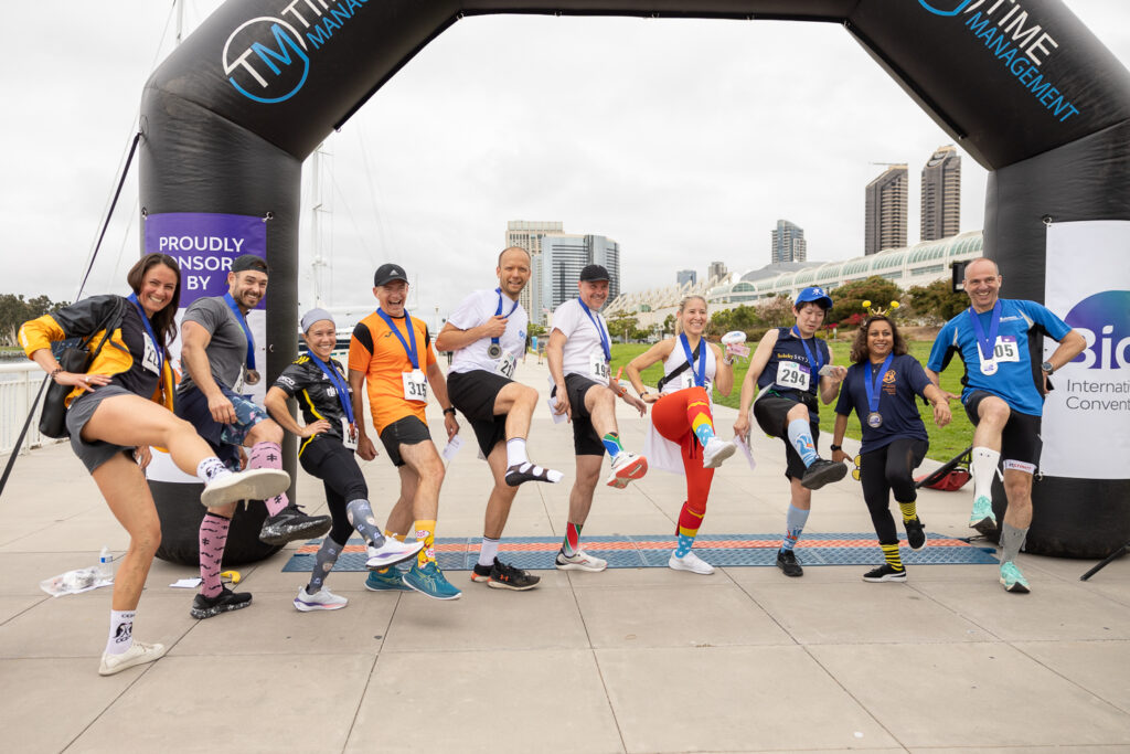 A group of people in athletic clothing pose playfully, lifting one leg each, under a finish line arch at an outdoor event. They are wearing race numbers and smiling, with a cityscape in the background.