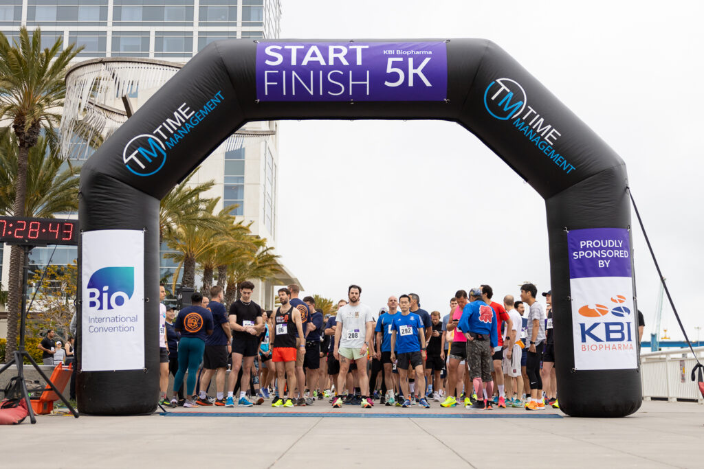 Runners gather at the starting line under an inflatable arch for a 5K race sponsored by KBI Biopharma and Bio International Convention. A timer shows 00:28:43 in the background.