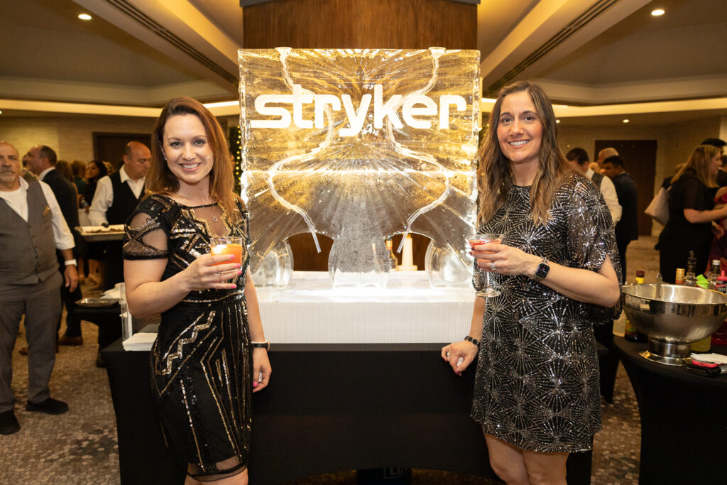 Two women in black dresses stand smiling, each holding a drink in front of an ice sculpture with the word "Stryker" engraved on it. The setting appears to be a formal indoor event with people mingling in the background.