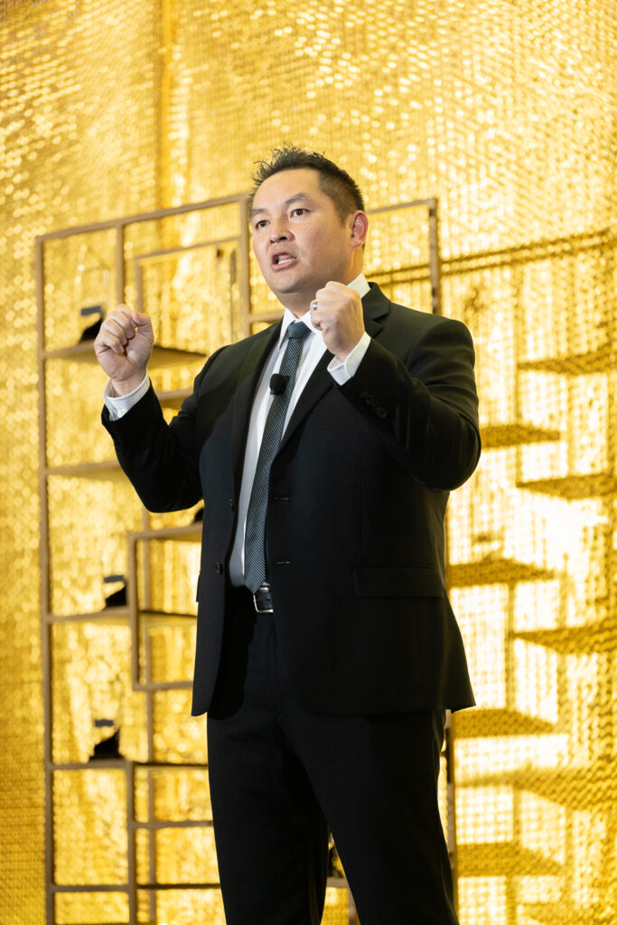 A man in a black suit and tie is speaking passionately on stage, gesturing with his fists raised. He stands in front of a shimmering gold background and a wireframe display shelf. He is wearing a microphone headset.