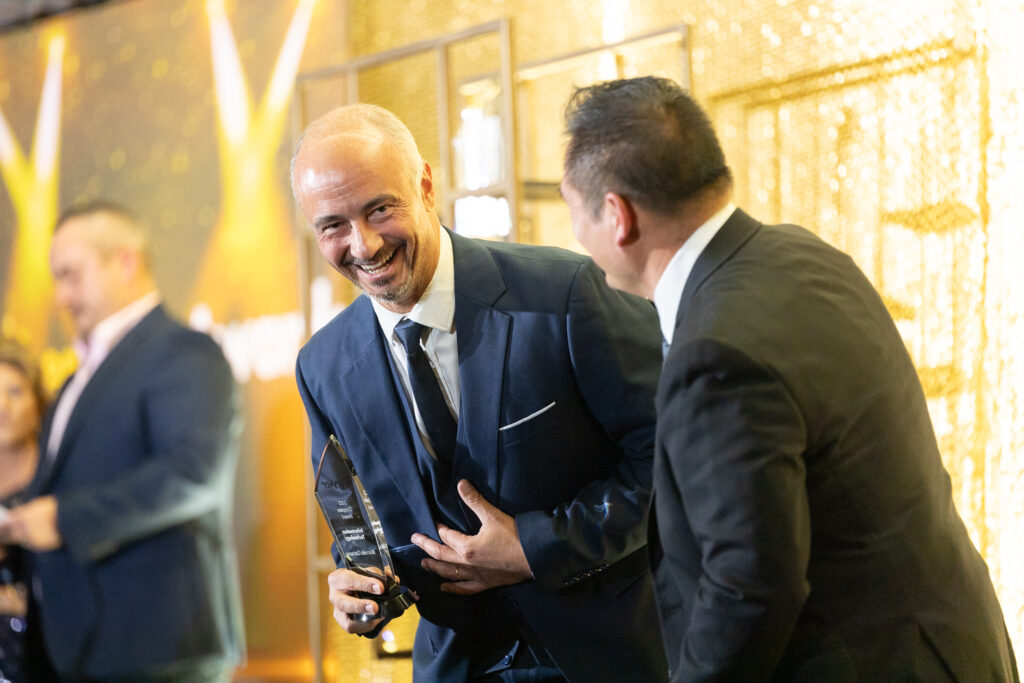 A man in a suit proudly holds a trophy while smiling and looking at another man who seems to be congratulating him. The background features a golden, shimmering backdrop and blurred figures.